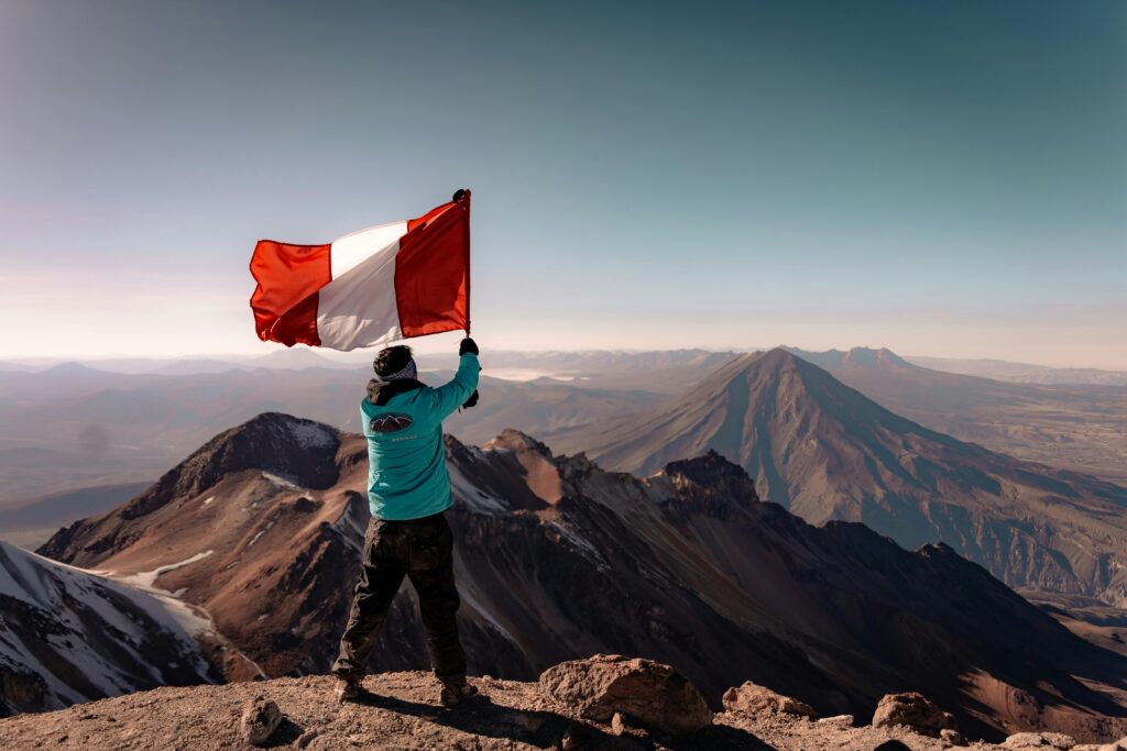 pexels-photo-27112858-27112858 A climber waves the Peruvian flag on a mountain peak with a breathtaking view of the Andes.
